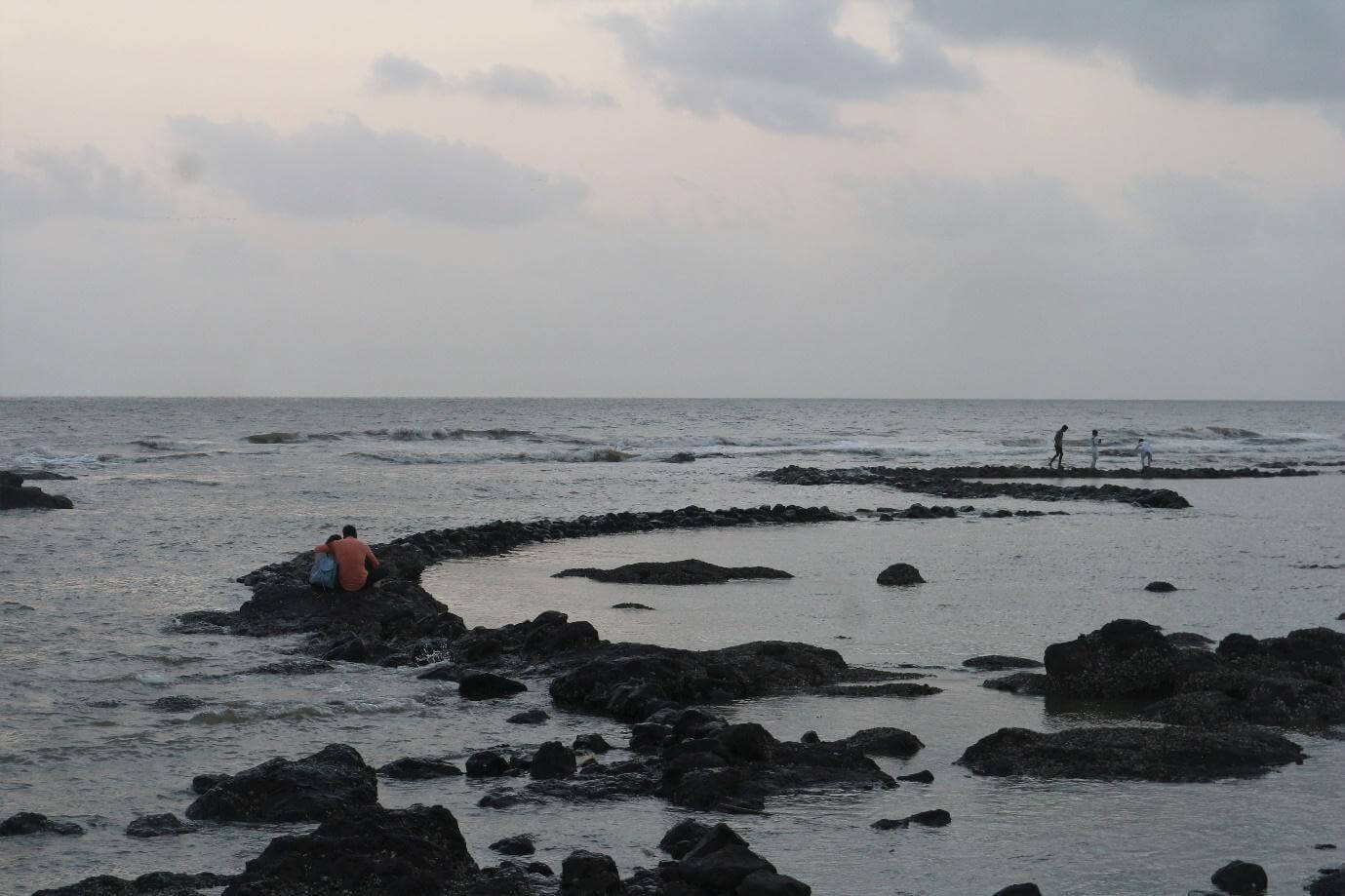 A man and a woman sit in a close embrace at the beginning of a trail of rocks leading farther into the sea